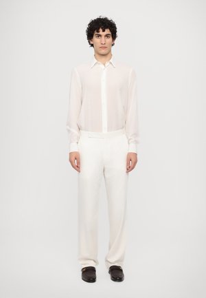 Hvid, skræddersyet skjorte med lange ærmer og knapper, parret med hvide bukser. Stoffet virker let og glat, suppleret med brune loafers.