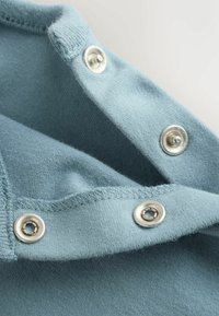 Gros plan d'un tissu bleu clair avec trois boutons-pression argentés, deux ouverts et un fermé, montrant le détail des coutures sur les bords.