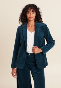 Blazer in corda di cotone verde acqua con revers e maniche lunghe, abbinato a pantaloni coordinati, indossato sopra una maglietta bianca a scollo a V.