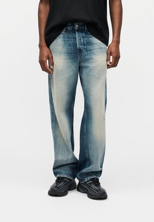 Osoba nosiaca vyblednuté modré široké rifle s jemným opotrebovaním a čierne športové topánky, stojaca pred jednoduchým bielym pozadím.