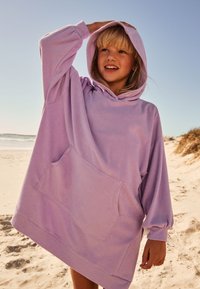Lila übergroßer Hoodie mit einer Fronttasche, weicher Textur, langen Ärmeln, verstellbarer Kordelzugkapuze, getragen an einem sandigen Strand.