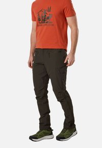 Homme portant un t-shirt orange avec un motif montagne et élan, un pantalon de randonnée vert foncé avec des poches zippées et des chaussures de trail vertes.