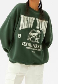 Zelená oversized mikina s bílým nápisem "NEW YORK" a grafikou psa, s žebrovými manžetami a zaobleným spodním lemem.