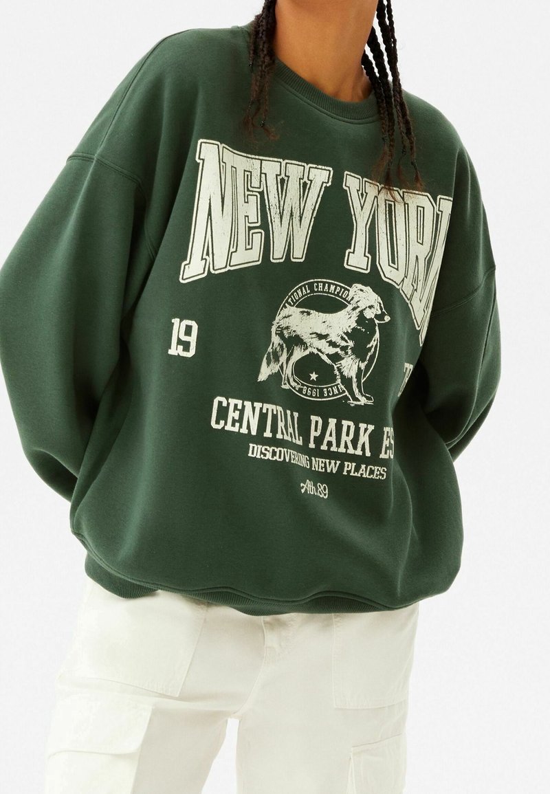 Zelená oversized mikina s bílým nápisem "NEW YORK" a grafikou psa, s žebrovými manžetami a zaobleným spodním lemem.