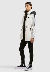 Kvinde iført hvid og sort hættetrøje med snore, sorte leggings og sorte høje sneakers, stående i profil på grå baggrund.