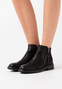 Botas de tobillo de cuero negro con punta redondeada, cremallera lateral, acabado texturizado y un tacón bajo en un fondo claro.