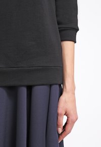 Femme portant un haut noir à manches longues et une jupe plissée foncée, avec son bras droit détendu le long du corps, sur fond blanc.