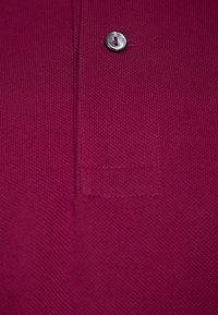 Camisa polo ajustada en tela de punto magenta oscuro, con un botón plateado en el collar y un detalle de parche sutil cerca del fondo.