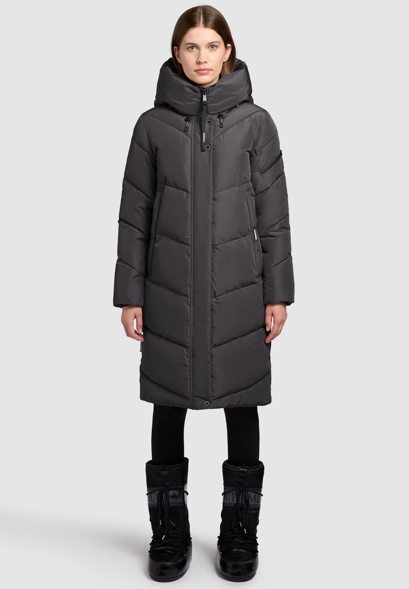 Geacă matlasată gri, cu guler înalt, fermoar, buzunare laterale și lungime până la genunchi; purtată cu cizme negre impermeabile.