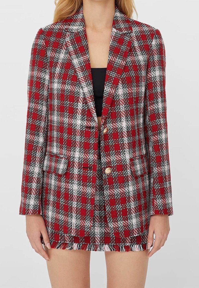 Femme portant un blazer à carreaux rouge et blanc avec des boutons dorés sur un top noir, assorti à une jupe courte à franges.