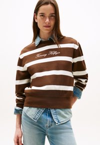 Felpa a righe marroni e bianche con la scritta "Tommy Hilfiger" ricamata in corsivo. Indossata sopra una camicia di jeans con toni blu.