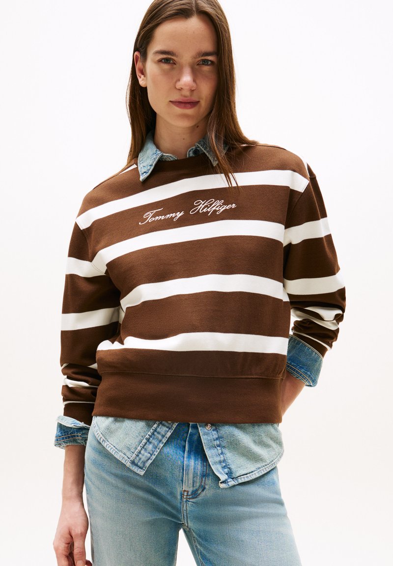 Felpa a righe marroni e bianche con la scritta "Tommy Hilfiger" ricamata in corsivo. Indossata sopra una camicia di jeans con toni blu.