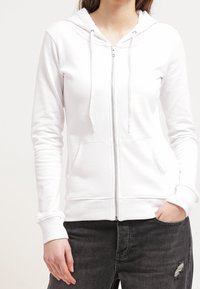 Sudadera blanca con cremallera y cordones, bolsillos tipo canguro, puños acanalados y textura suave, combinada con jeans negros rasgados.