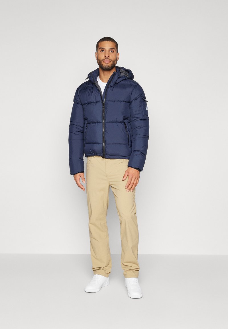 Marineblaue Steppjacke mit Kapuze, Reißverschluss und Seitentaschen, kombiniert mit beigen Hosen und weißen Sneakern. Glatte Textur, gesteppter Design.