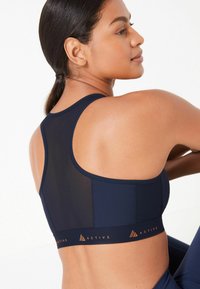 Marineblauwe sportbeha met meshpanelen, racerback ontwerp en oranje logo-band. Glad textuur en aansluitende pasvorm voor atletische ondersteuning.