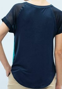 Navy t-shirt with short sheer sleeves, round neckline, and a curved hem. Features a solid back and a contrasting teal side panel with dots.