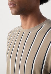 T-shirt beige à rayures avec de fines rayures verticales noires et blanches. Encolure ronde, texture douce en coton, coupe décontractée avec manches courtes.