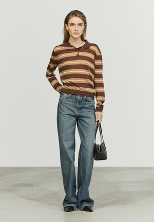 Ung kvinde iført brunstribet langærmet poloshirt, løse blå jeans, der holder en lille sort håndtaske, stående mod en ensartet lys baggrund.