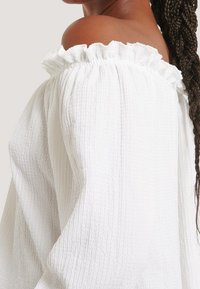 Weißes Off-Shoulder-Oberteil mit gerafftem Ausschnitt, strukturiertem Stoff und langen Ärmeln. Die Webart weist ein dezentes, erhabenes Muster auf.