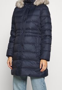 Abrigo acolchado azul marino con capucha desmontable forrada de piel, patrón acolchado, botones frontales y mangas largas. Presenta un ajuste holgado y bolsillos laterales.