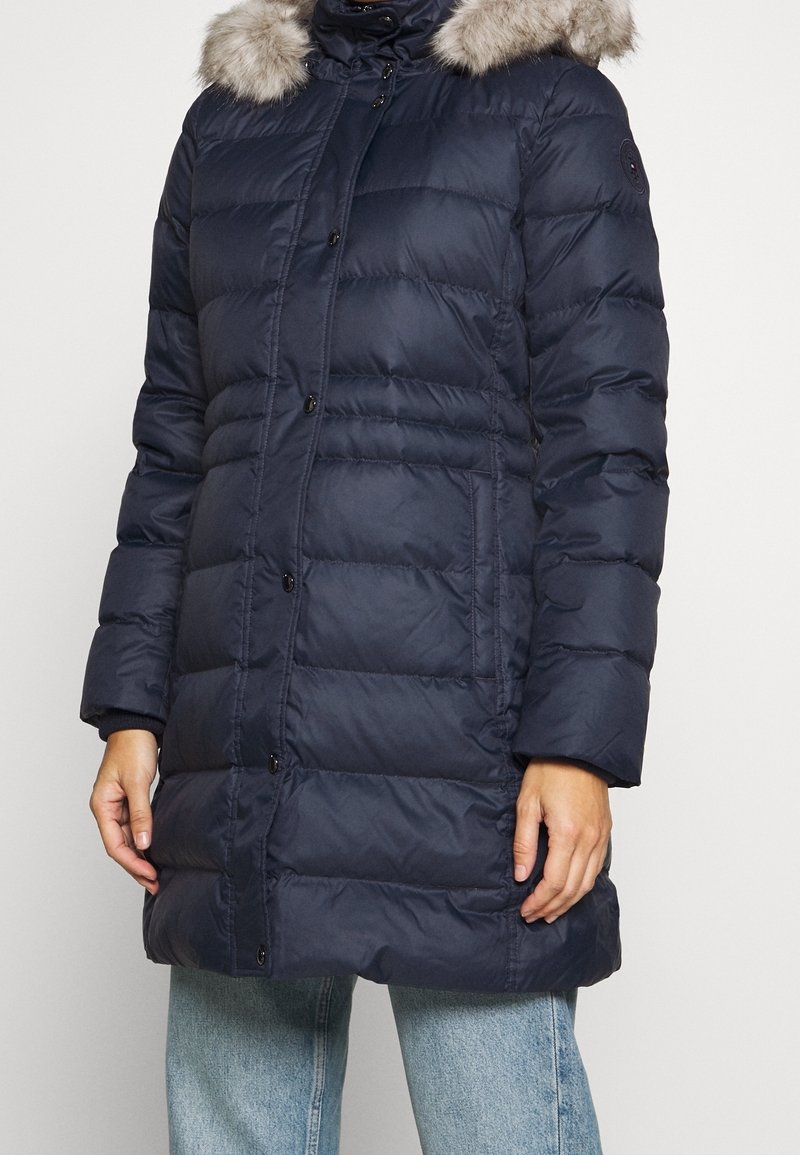 Abrigo acolchado azul marino con capucha desmontable forrada de piel, patrón acolchado, botones frontales y mangas largas. Presenta un ajuste holgado y bolsillos laterales.