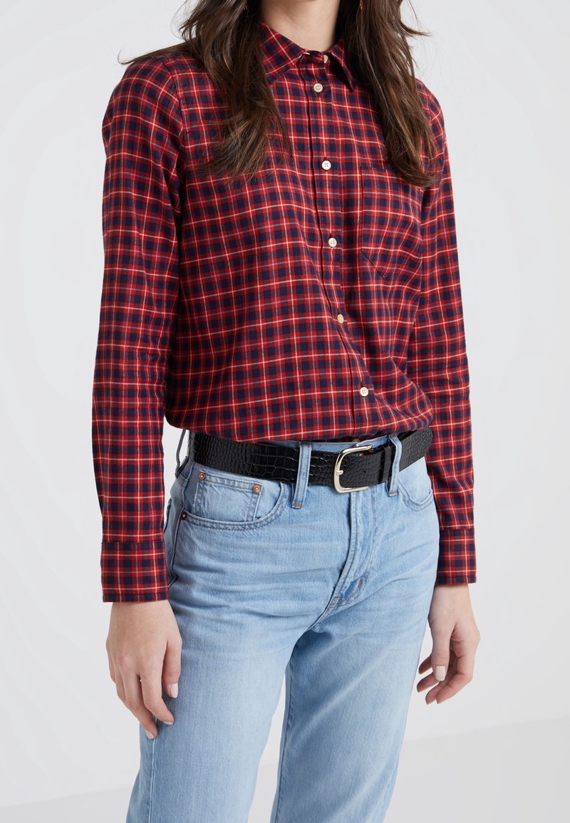 Femme portant une chemise à carreaux rouge et bleu marine boutonnée, rentrée dans un jean bleu clair avec une ceinture noire texturée.