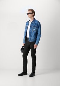 Jeansjacke in Hellblau, schwarze Hose, schwarze Schuhe, weißes T-Shirt und quadratische schwarze Sonnenbrille. Als Accessoire dient eine kleine schwarze Tasche.