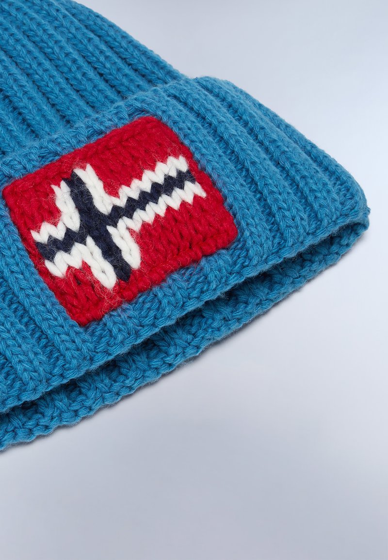 Blauer gestrickter Beanie mit einem roten Patch, das ein weißes und marineblaues Design zeigt. Gerippte Textur, umgeschlagener Bund und dicke Garnkonstruktion.