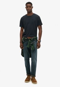 Mand iført sort t-shirt, blå jeans, brune støvler, brunt ur og en grønternet skjorte bundet om livet, står mod en hvid baggrund.