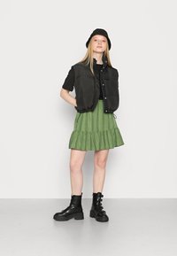 Colete puff preto sobre uma camiseta preta de mangas curtas, saia plissada verde com riscas verticais e botas pretas de amarrar com fivelas.