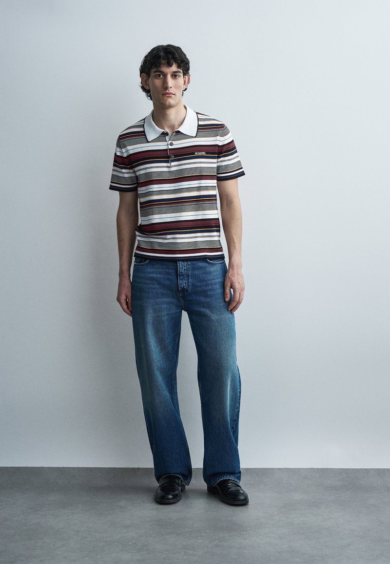 Young man standing against gray wall wearing striped polo shirt, loose blue jeans, and black shoes, facing forward with neutral expression.