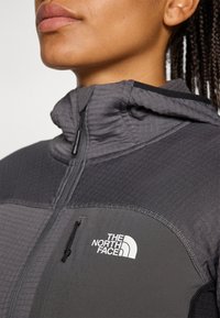 Lähedane vaade naise kaelale ja lõuale, kes kannab halli North Face'i jope, millel on nähtav lukuga tasku ja logo.