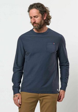 Homme aux cheveux bouclés et à la barbe portant une chemise bleu marine à manches longues avec une poche poitrine et un pantalon marron, regardant vers le bas à sa gauche.