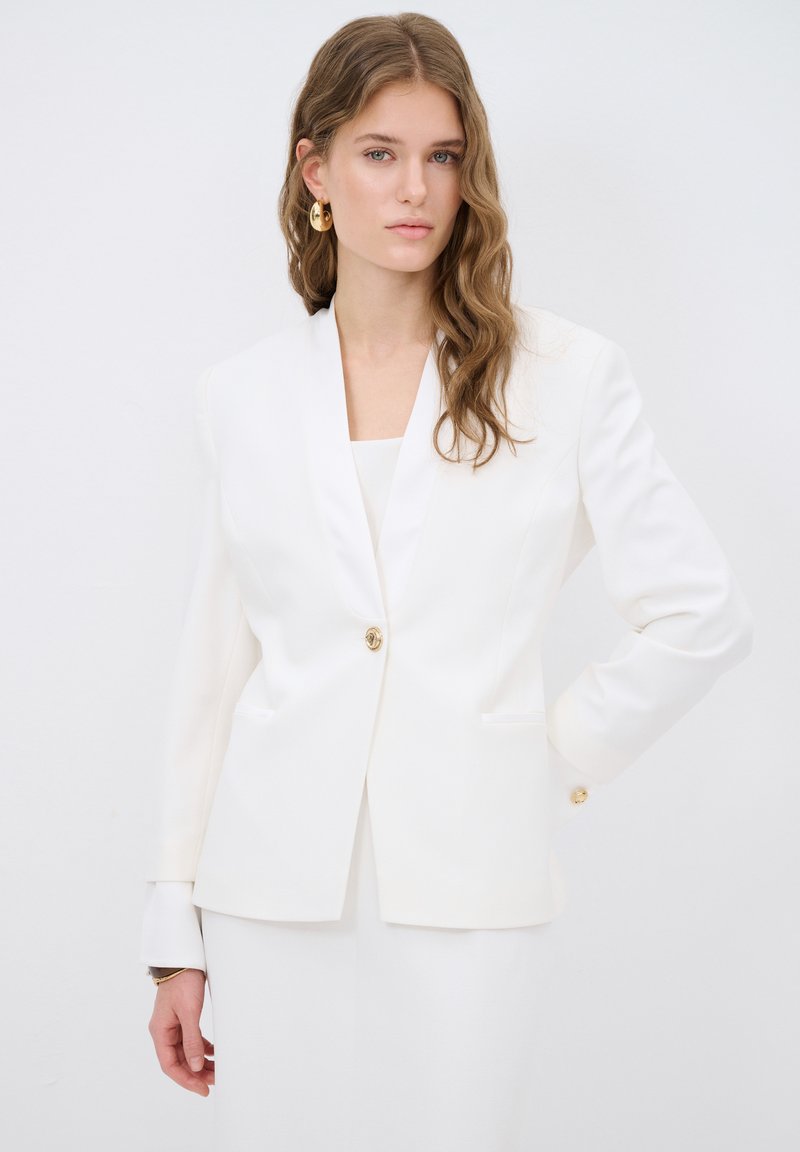 White blazer with a notched collar and single gold button. Tailored fit with two front pockets and long sleeves, paired with a white top.