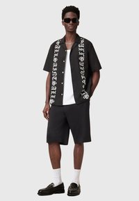 Chemise noire à manches courtes avec un design de texte contrasté en blanc. Assortie à un short noir et des mocassins, accessoirisée avec des lunettes de soleil et des chaussettes blanches.