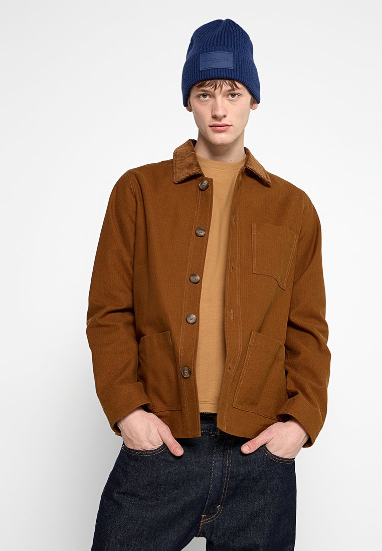 Brown corduroy jacket with a collar, front button closure, two side pockets, and a single chest pocket. Worn over a tan shirt and dark jeans. Blue beanie.