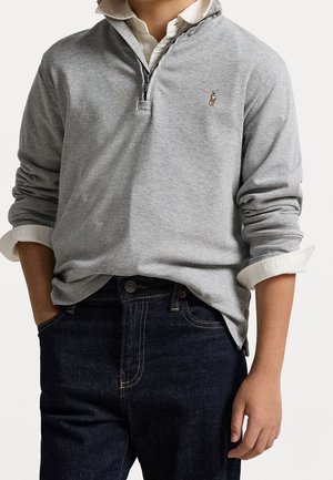 Persoon draagt een lichtgrijze pullover met een kwartritssluiting over een wit overhemd met opgerolde mouwen, gecombineerd met donkerblauwe spijkerbroek.