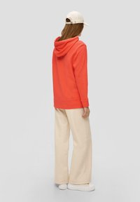 Korallfärgad hoodie med dragsko, avslappnad passform, ribbade ärmslut och nederkant. Kombinationen med beige vida byxor och vita sneakers, samt en beige keps.