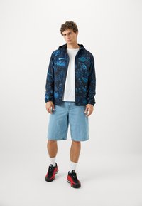 Sort hættejakke med blåt abstrakt mønster, hvid t-shirt, lyseblå denim shorts og sort/røde sneakers med hvide sokker.