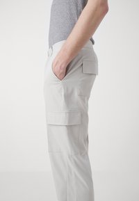 Pantalones cargo gris claro con un diseño ajustado, que presentan bolsillos laterales y un bolsillo parcheado en el muslo, fabricados con una tela suave.