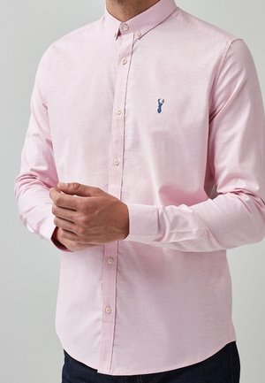 Homme portant une chemise à manches longues rose clair boutonnée jusqu'en haut avec un petit logo de cerf bleu foncé sur la poitrine gauche, les mains jointes devant lui.