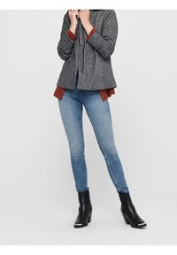 Grijze gebreide jas met een capuchon, voorzien van een rits en drukknoopsluiting, gedragen over een rood langemouwen shirt. Gecombineerd met lichtblauwe skinny jeans en zwarte hoge laarzen.