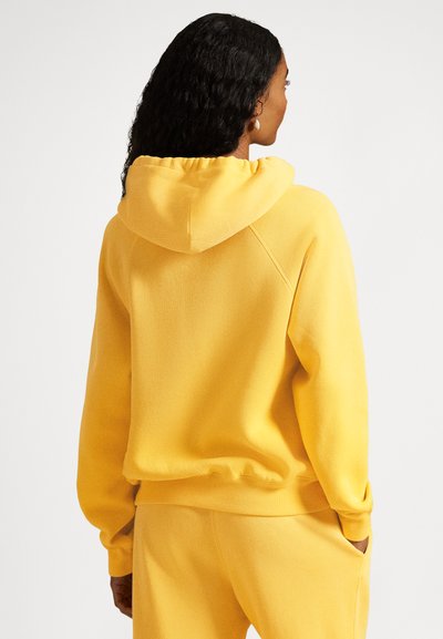 Rumena pulover s kapuco z sproščenim krojem, rebrastimi manšetami in spodnjim robom. Mehak tekstura tkanine, spredaj ima žep v stilu kenguruja.