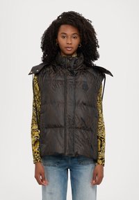 Gilet imbottito marrone con finitura lucida, colletto alto e dettagli con bottoni a pressione. Presenta un logo impresso e si abbina a una camicia a fantasie.