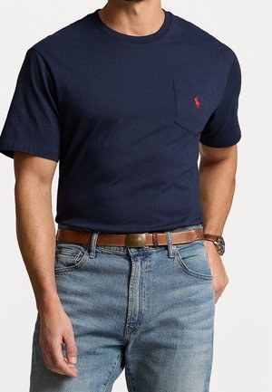 Homme portant un t-shirt bleu marine à manches courtes avec une poche poitrine et un logo rouge, un jean bleu clair, une ceinture en cuir marron et une montre-bracelet.