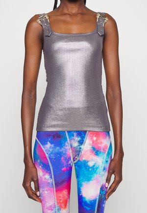 Vrouw met een glanzende grijze mouwloze top met versierde schouderbanden en kleurrijke leggings met een galaxypatroon, staand tegen een effen achtergrond.