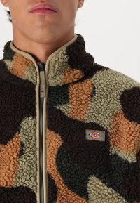 Giacca in fleece con un motivo camouflage in marrone, verde e sabbia. Presenta una chiusura con zip e una patch del logo Dickies sul lato sinistro.