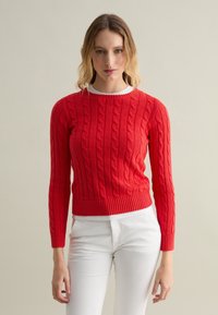 Maglione rosso a maglia cable con maniche lunghe, collo rotondo e orlo a coste, abbinato a pantaloni bianchi. Motivo testurizzato di trecce intrecciate.