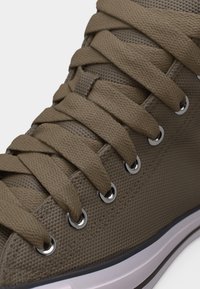 Krupni plan smeđe tenisice s teksturom, širokim platnenim vezicama provučenim kroz srebrne metalne rupice i bijelim gumenim potplatom.