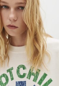 Jonge vrouw met lang blond haar, gekleed in een witte sweatshirt met versleten groene en blauwe tekst op een neutrale achtergrond.